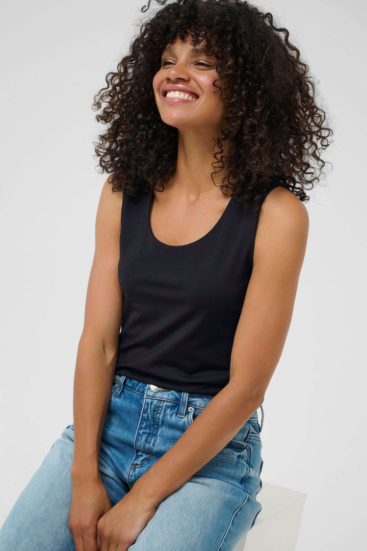 Femme souriante portant un débardeur noir et un jean, assise sur un tabouret, les cheveux bouclés et détendus.