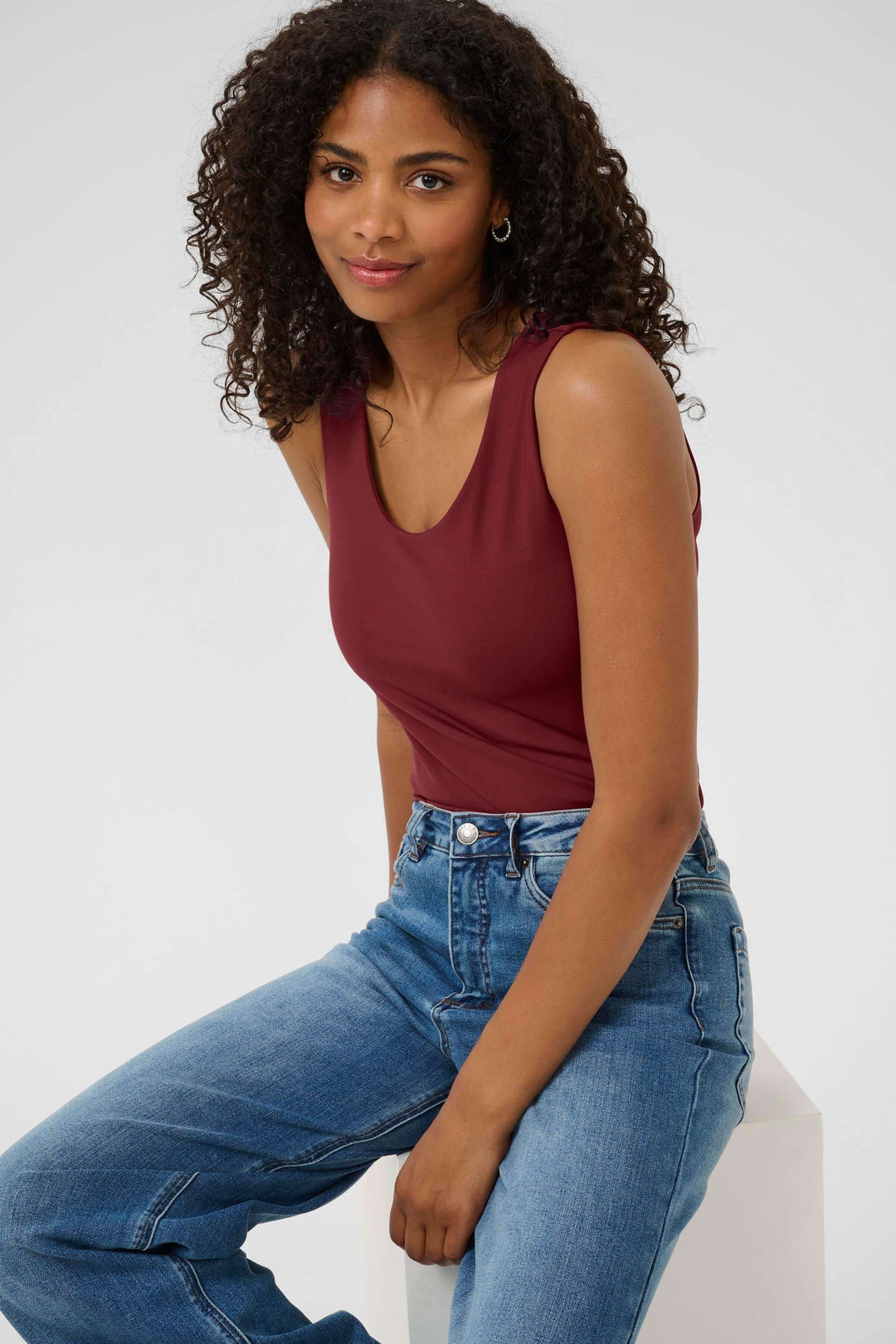 Femme portant un débardeur bordeaux avec jean, posant assise, sur fond gris clair.