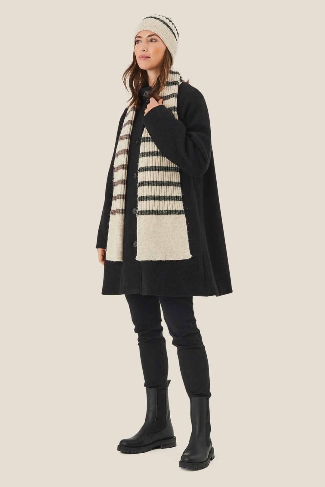 Femme portant une tuque lignée et une écharpe rayée, vêtue d'un manteau noir sur fond crème.