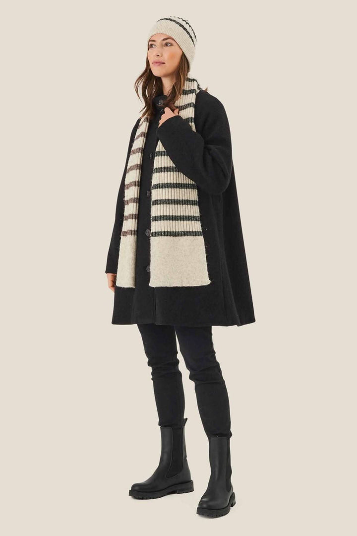 Femme portant une tuque lignée et une écharpe rayée, vêtue d'un manteau noir sur fond crème.