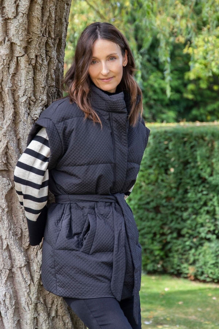 Modèle portant un gilet matelassé noir Tahani avec ceinture, posé contre un arbre, entouré d'une verdure.