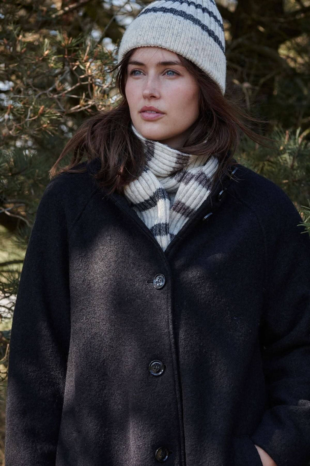 Femme portant un bonnet à rayures gris et blanc, écharpe à carreaux, et manteau noir en pleine nature.