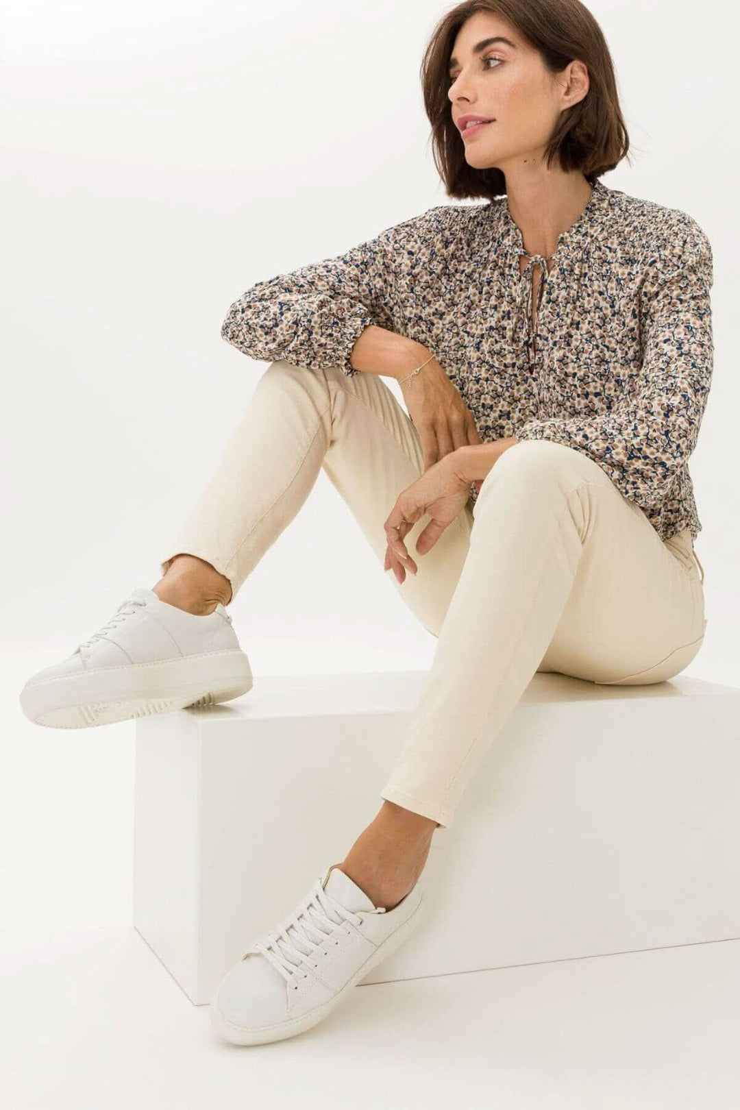 Femme portant le pantalon ANA avec blouse à motifs, assise sur une plateforme, chaussures blanches élégantes.