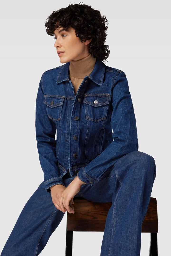 Femme portant une veste en jean bleu avec manches longues et poches, assise sur une chaise en bois.