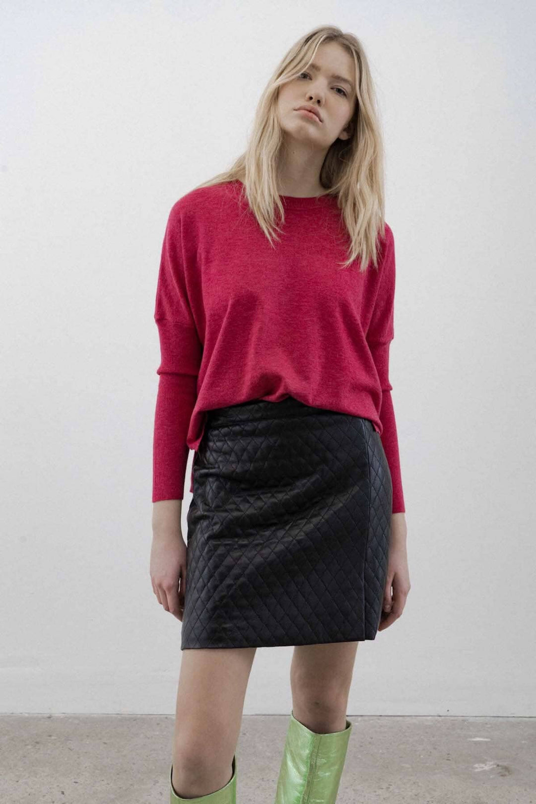 Femme portant une jupe courte noire à motifs de losanges et un pull rose, stylée avec des bottes vertes.