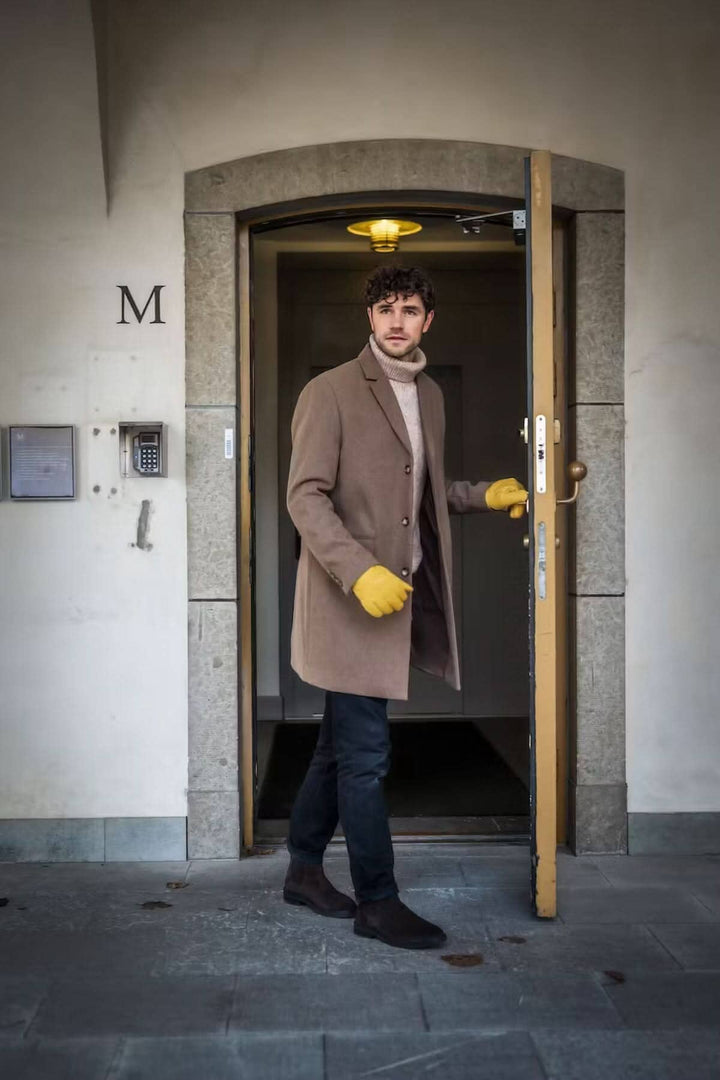 Homme sortant par une porte, portant un manteau beige et des gants jaunes, élégant et stylé.