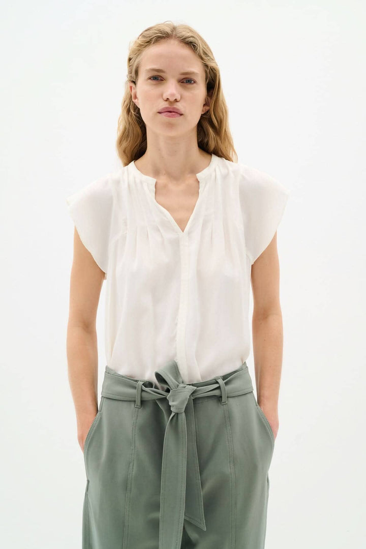 Blouse Cidenza blanche légère avec encolure plissée et manches volantes, portée avec un pantalon vert.
