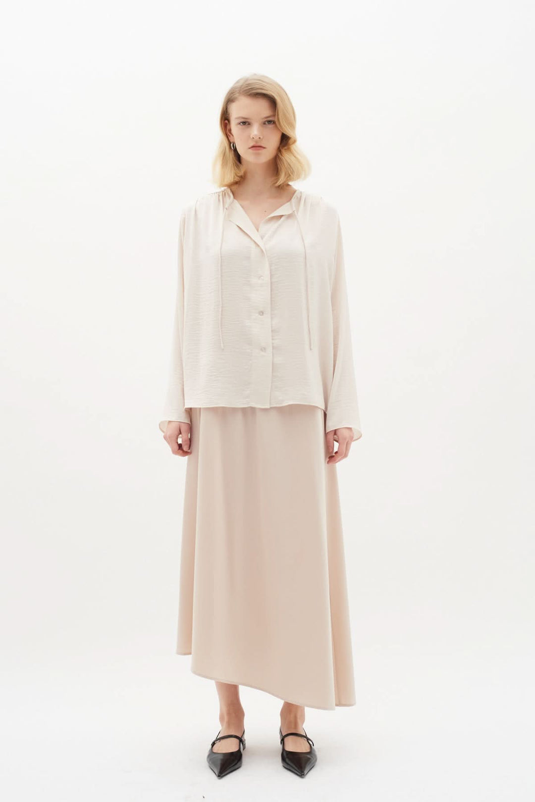 Femme portant une blouse Mato à manches longues avec encolure nouée, associée à une jupe longue beige.