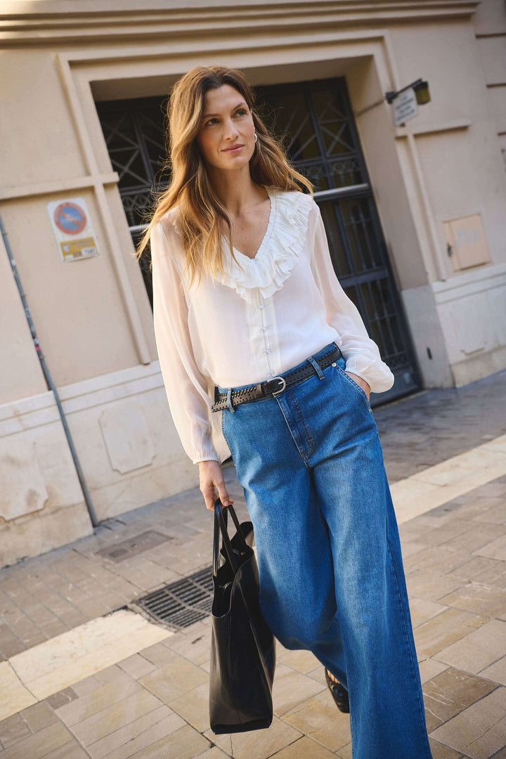 Femme portant une blouse fluide blanche avec volants et un pantalon en denim large dans une rue urbaine.