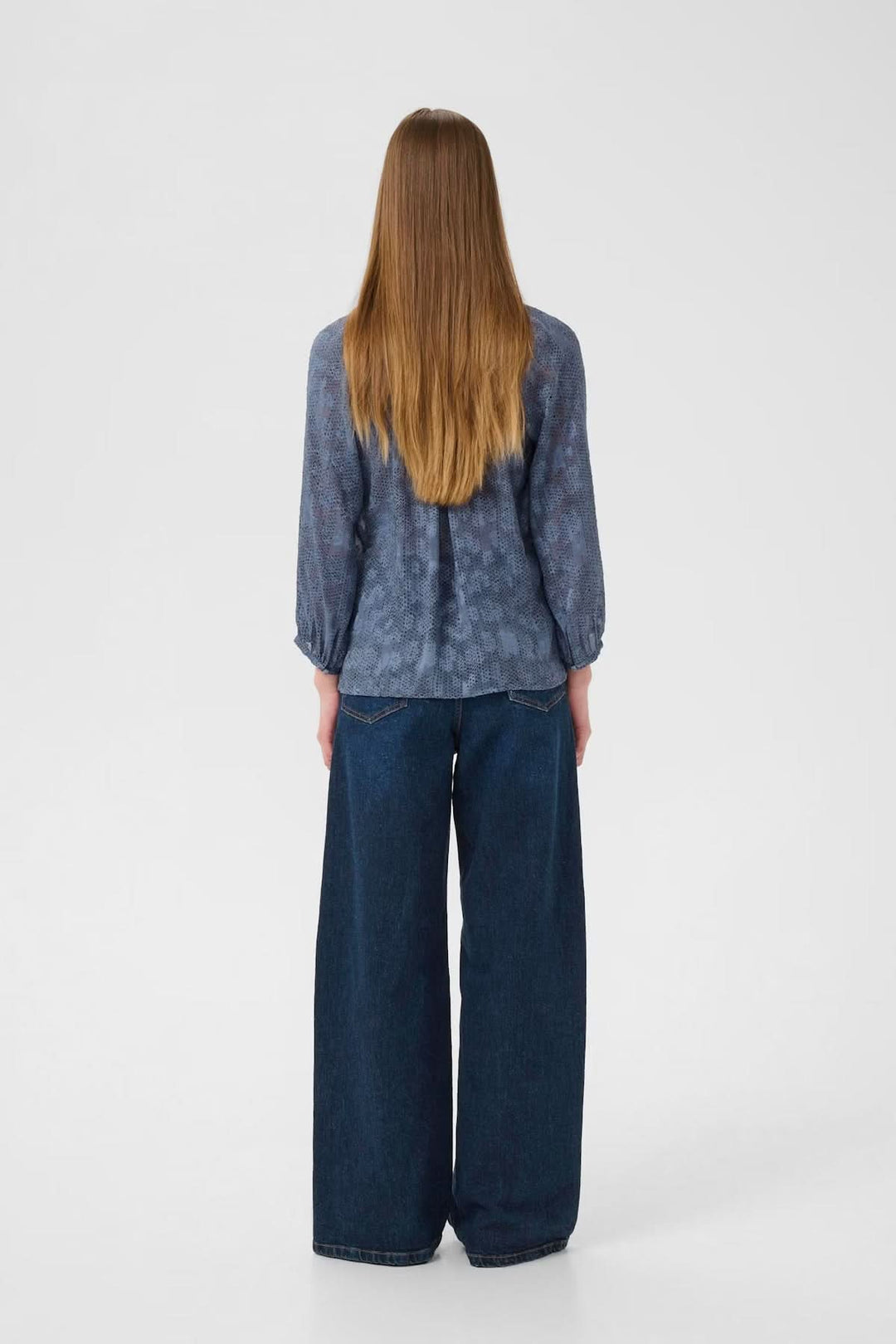 Femme vue de dos portant une blouse à col V et manches 3/4 avec un pantalon large en denim.