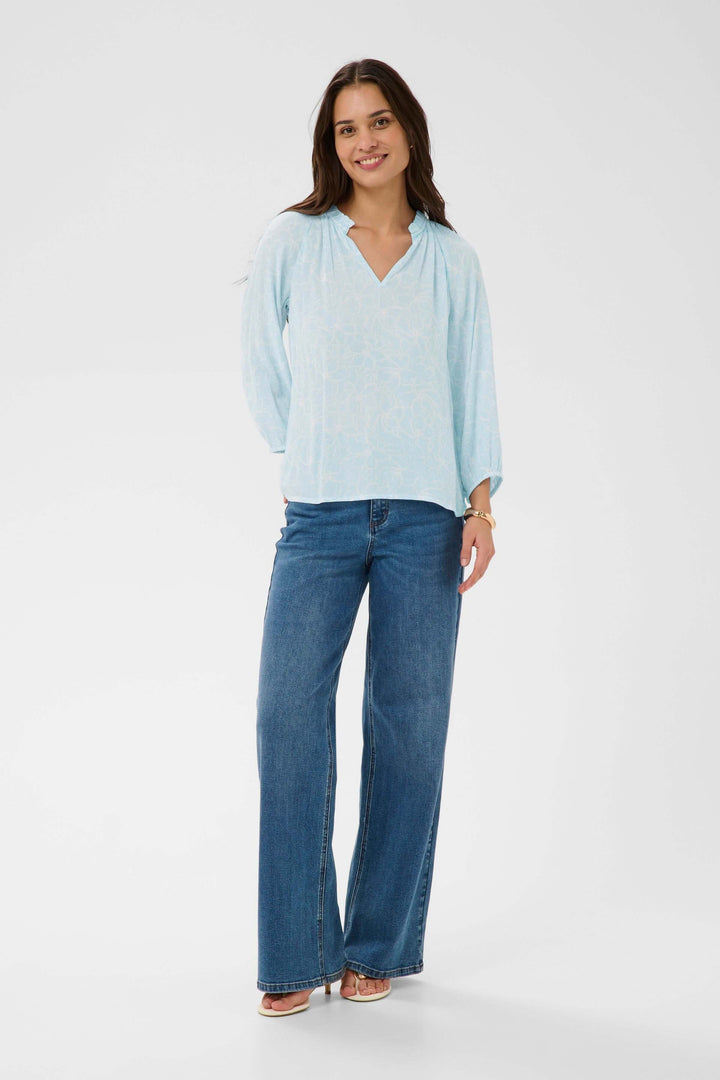 Femme portant une blouse fluide à imprimé floral discret avec encolure en V et manches trois-quarts, style décontracté.