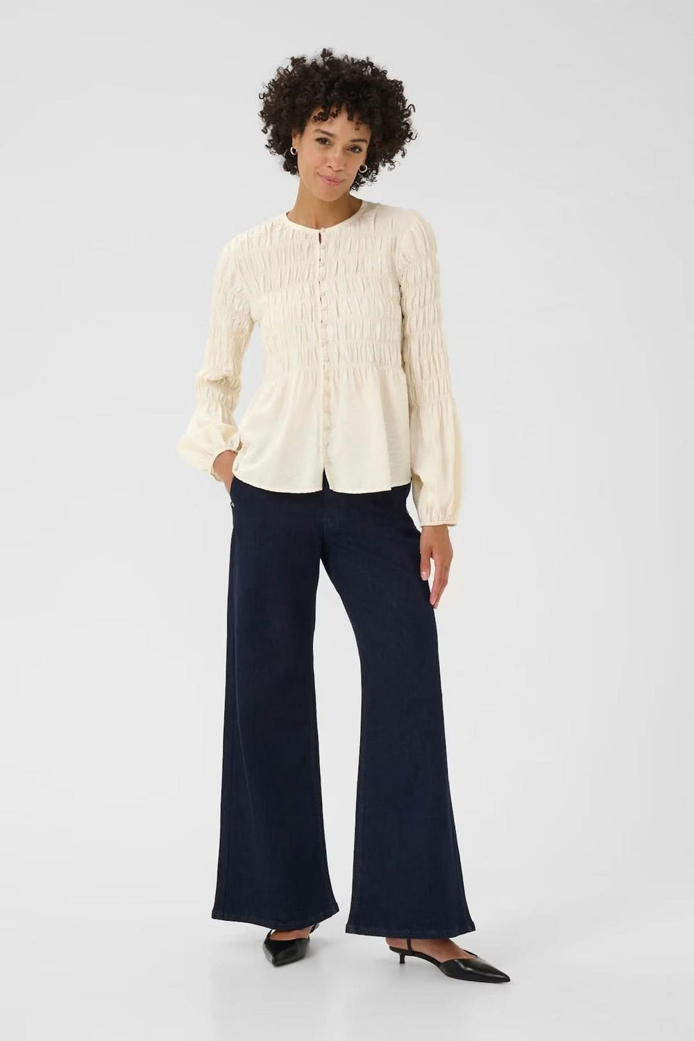 Femme portant une blouse à fronces avec encolure ronde, coupe fluide et détails texturés, associée à un pantalon large pour un look chic.