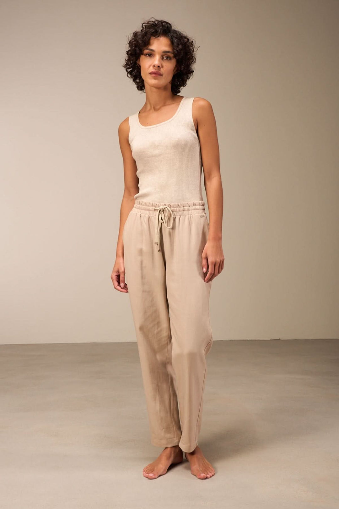 Femme en camisole beige sans manches et pantalon léger, posant sur un fond neutre.