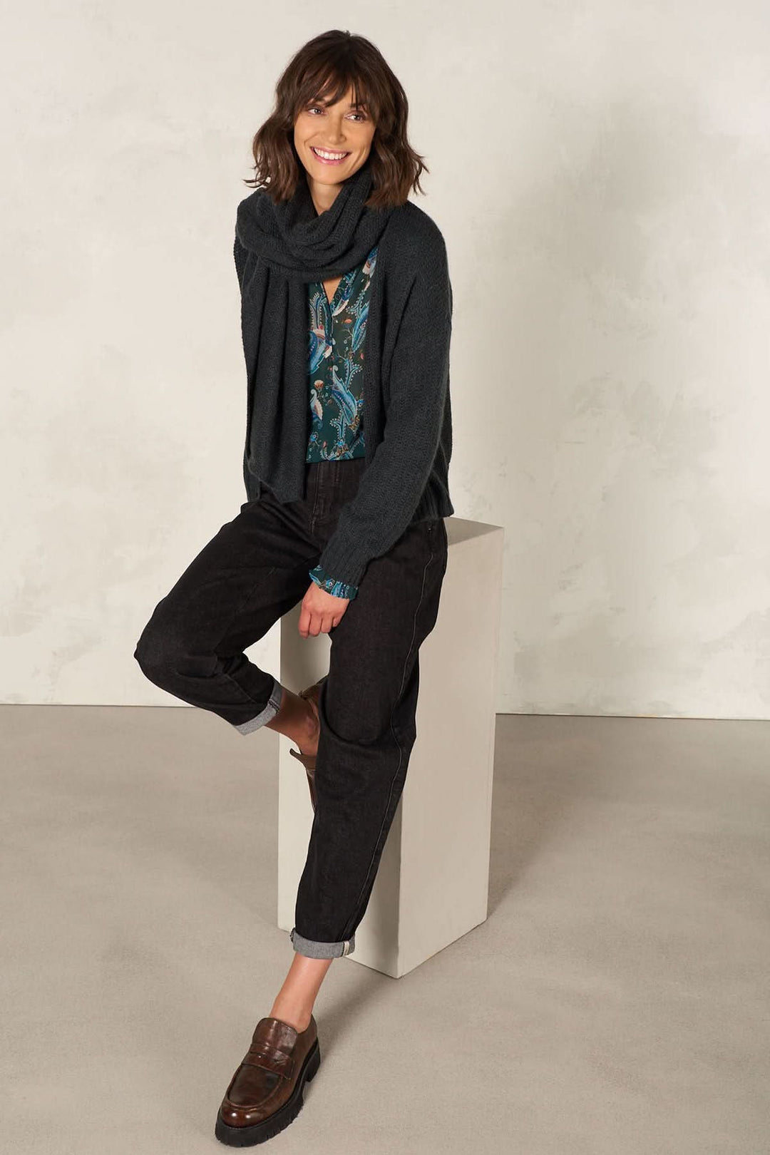 Femme souriante portant une cape foulard multifonctionnelle avec un pantalon noir sur un fond neutre.