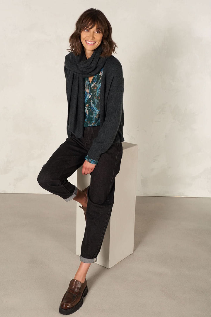 Femme souriante portant une cape foulard multifonctionnelle avec un pantalon noir sur un fond neutre.