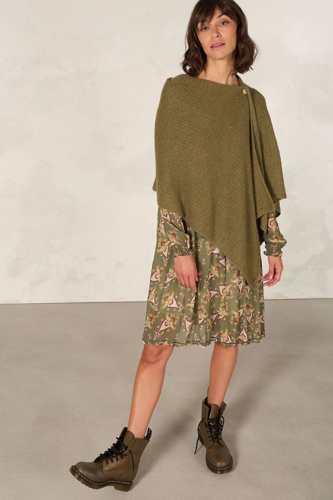 Femme portant une cape foulard tricotée verte, associée à une robe à motifs et des bottes, dans un décor minimaliste.