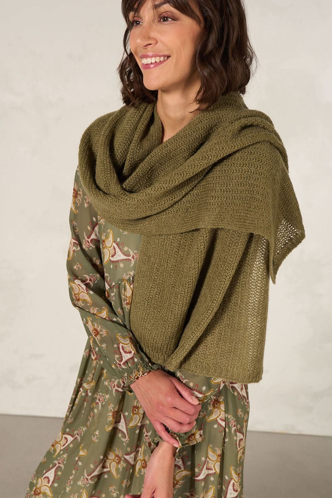 Femme souriante portant une cape en tricot multifonctionnelle de couleur olive sur une robe imprimée.