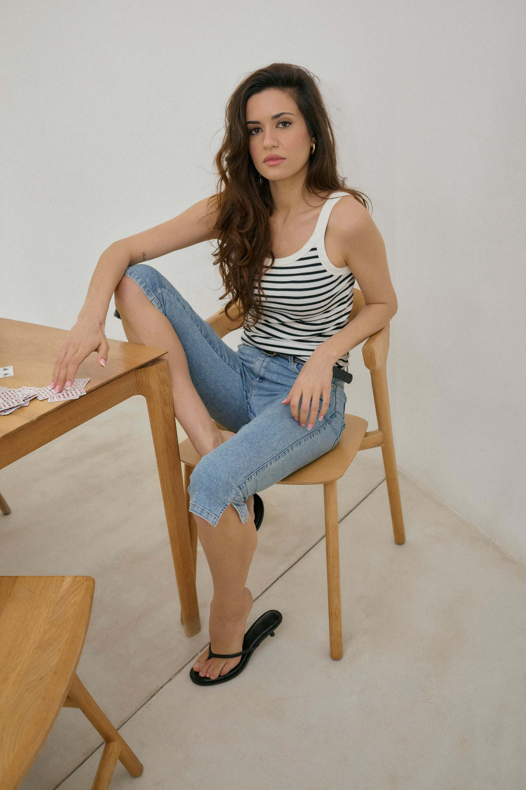 Femme assise sur une chaise en bois, portant un haut à rayures et un capri en denim sous le genou.