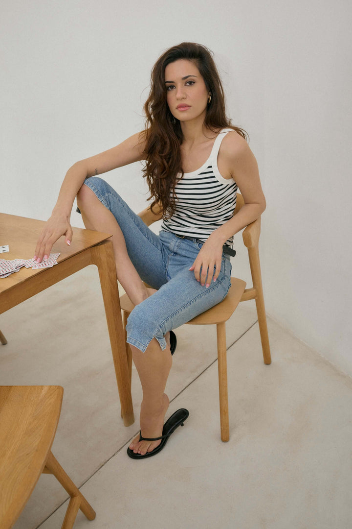 Femme assise sur une chaise en bois, portant un haut à rayures et un capri en denim sous le genou.