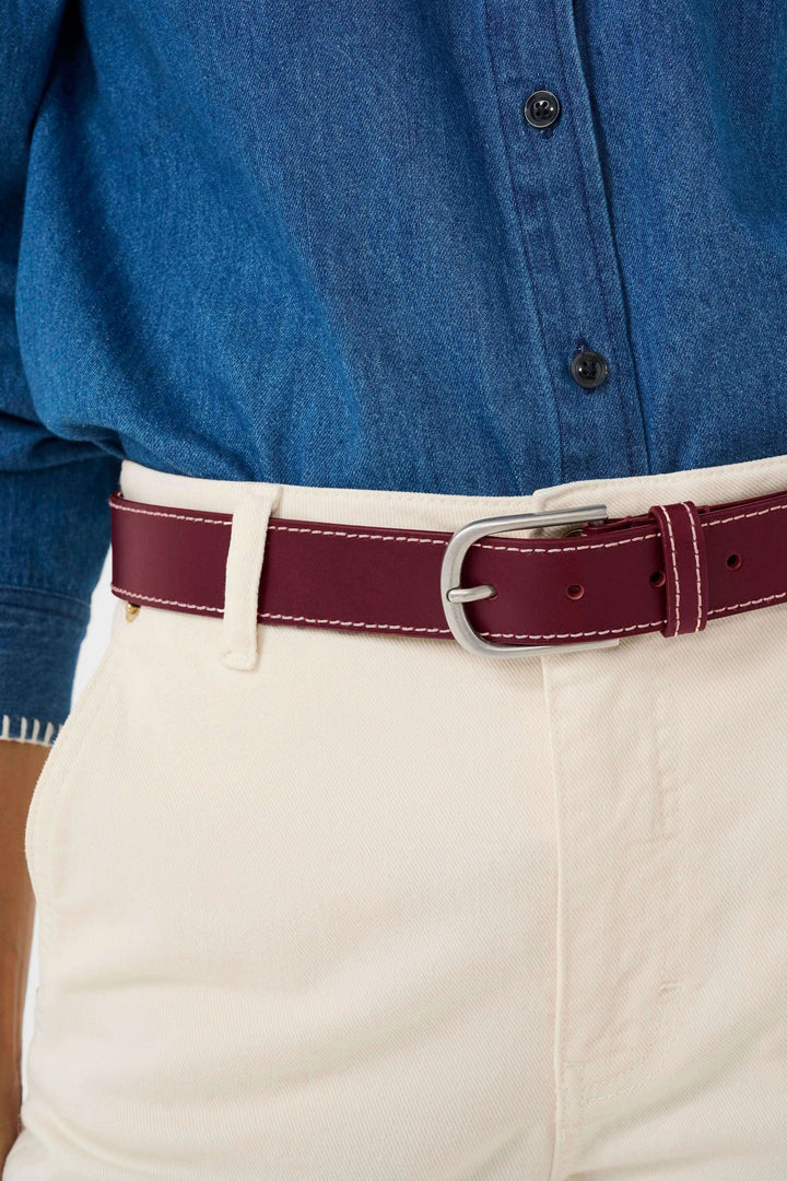 Ceinture Seyra en cuir véritable bordeaux avec surpiqûres contrastantes et boucle métallique classique, portée avec une chemise en jean et un pantalon beige.