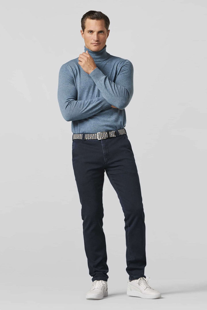 Homme en pull à col roulé bleu avec ceinture tressée, pantalon noir, chaussures blanches, posant avec assurance.