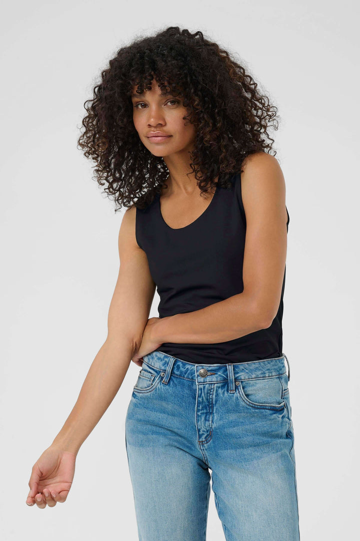 Person wearing black sleeveless top with curly hair, styled with blue jeans against a plain background.