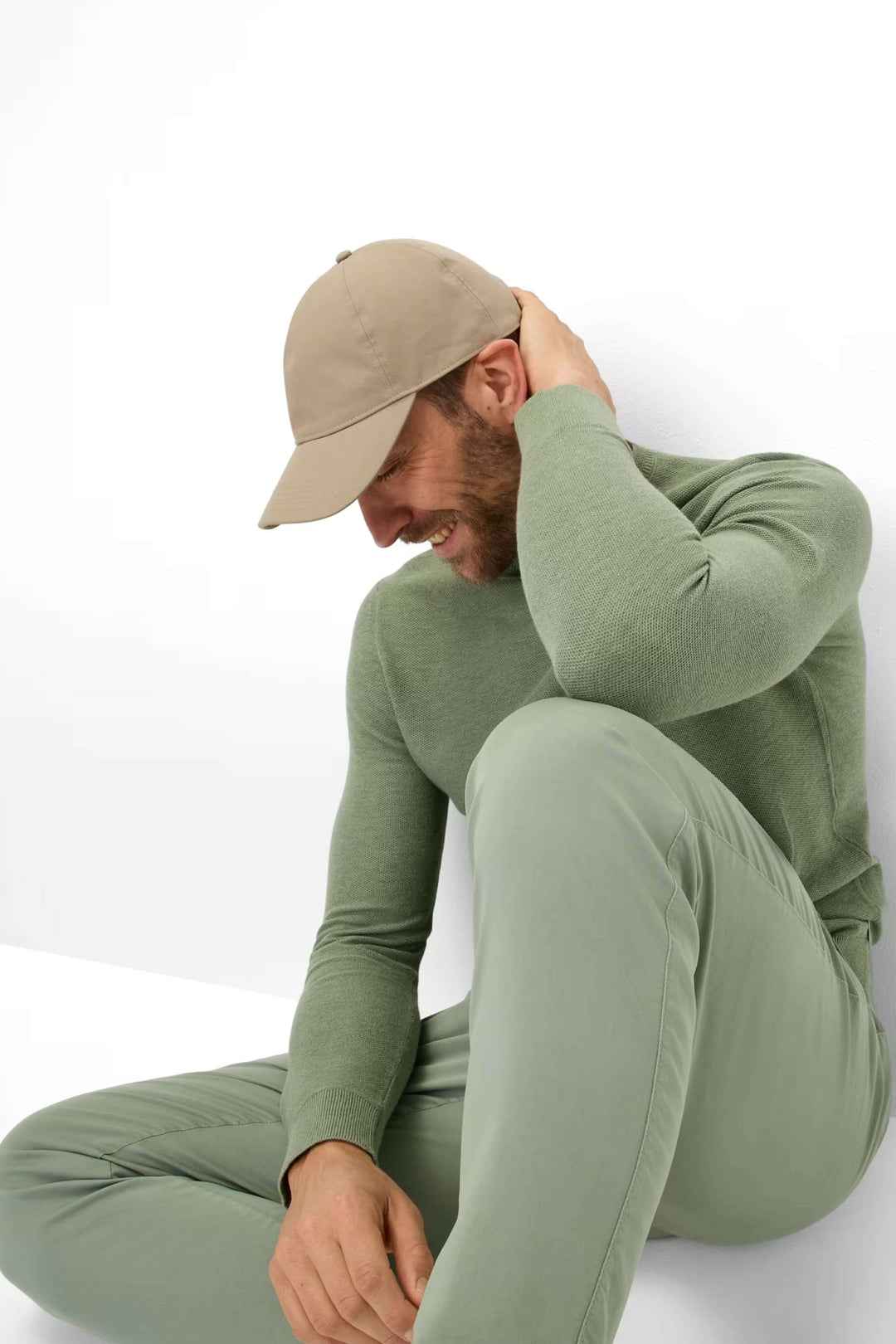Homme souriant portant un chandail vert et une casquette beige, assis sur le sol.