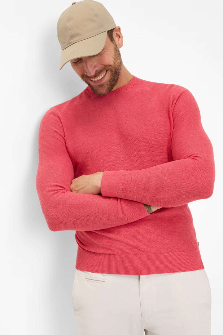 Homme souriant portant un chandail à col rond rose et une casquette beige, affichant un style décontracté.