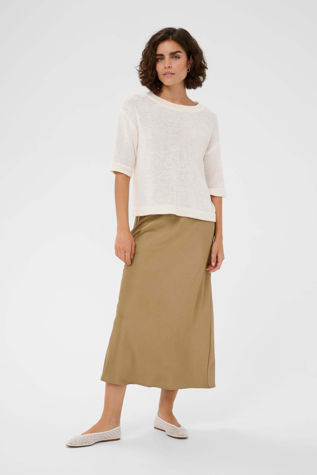 Femme portant le Chandail Sevita blanc, coupe droite, encolure ronde, manches courtes, sur une jupe longue beige, style contemporain.