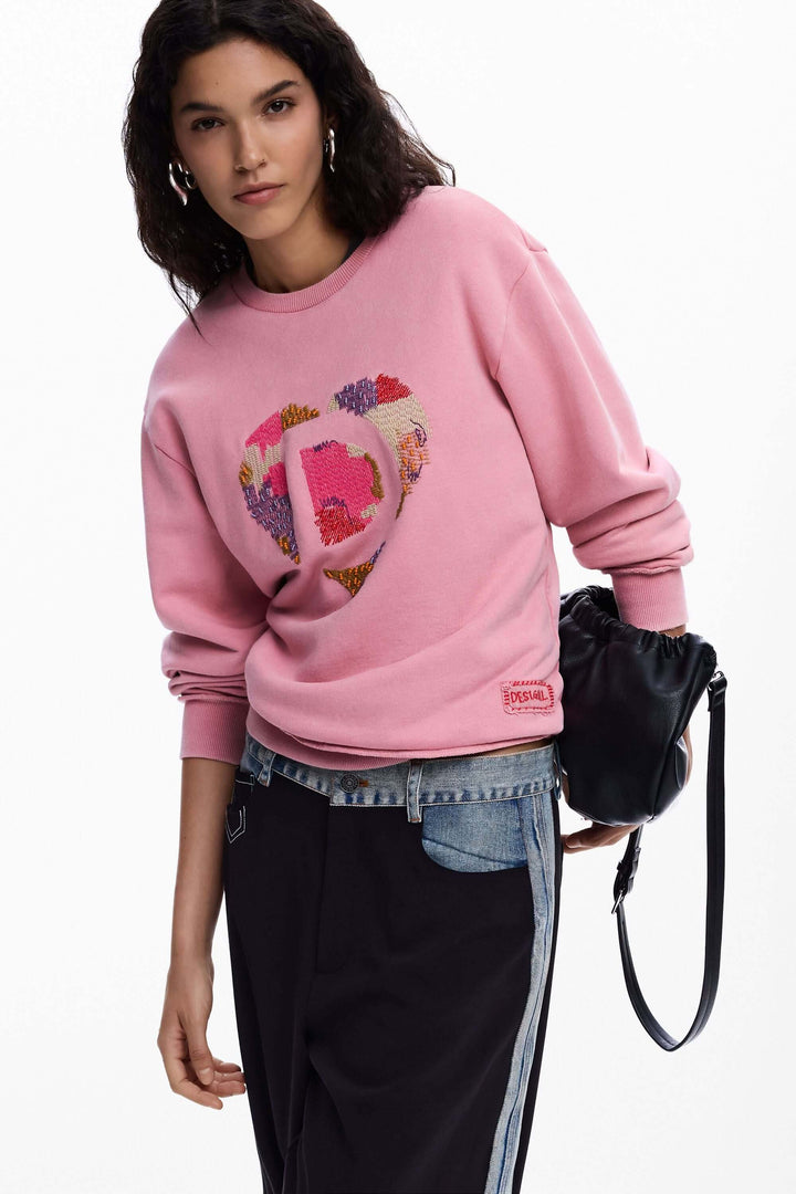 Femme portant un chandail rose avec logo et perles, manches longues, motif brodé graphique, coupe droite, style décontracté.