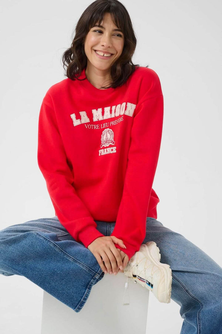 Chandail à col rond rouge avec inscription brodée, style décontracté universitaire, porté avec un jean.