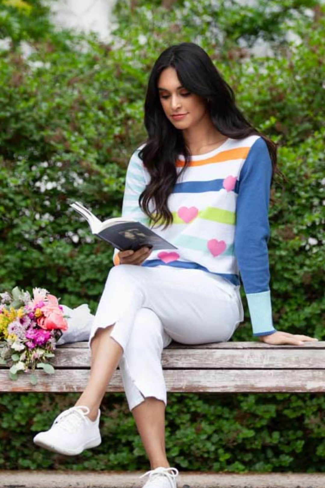 Femme lisant un livre sur un banc, portant un chandail à rayures colorées et cœurs, avec un bouquet de fleurs.