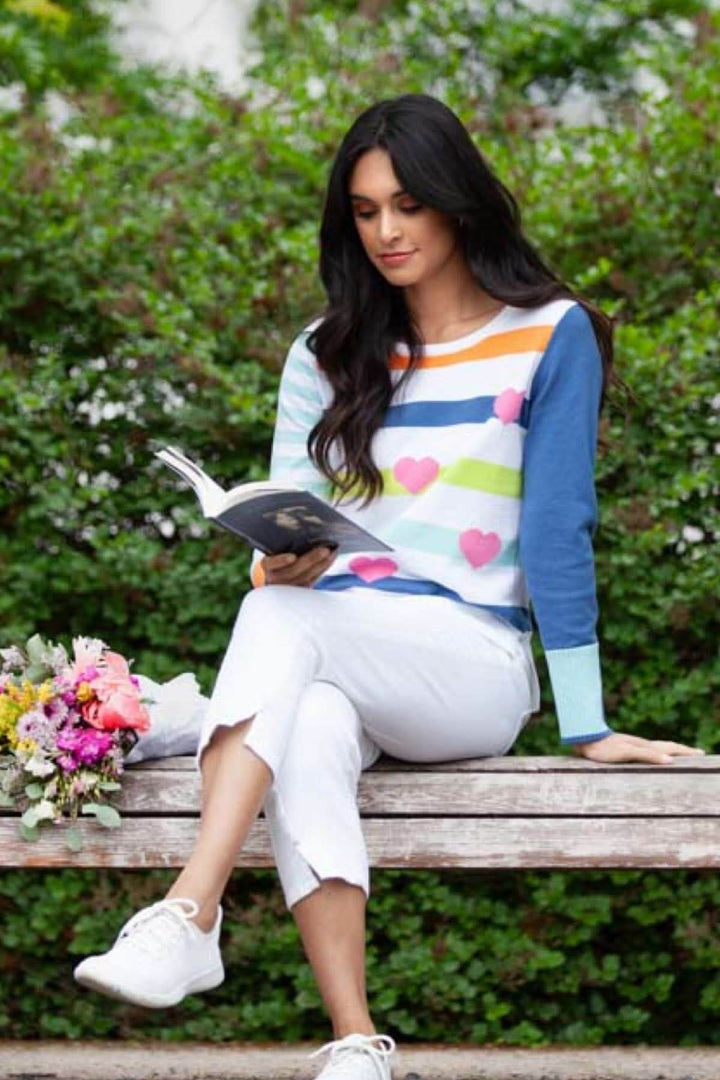 Femme lisant un livre sur un banc, portant un chandail à rayures colorées et cœurs, avec un bouquet de fleurs.