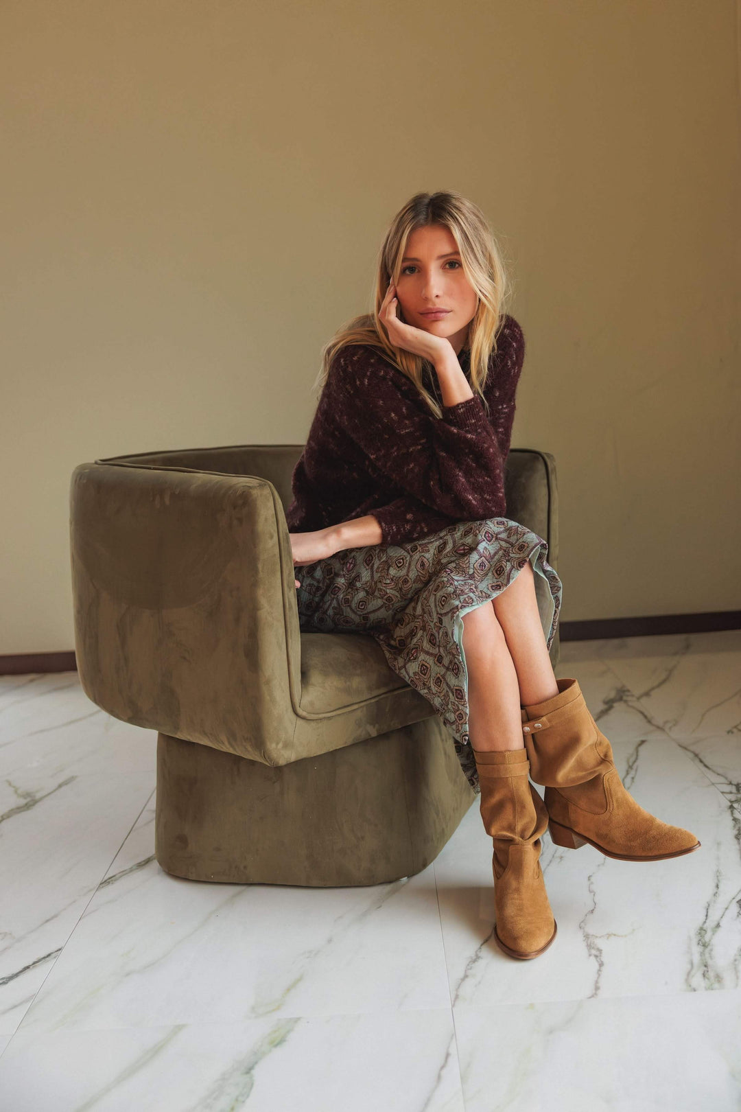 Femme assise dans un canapé olive, portant un chandail maille texturée et une jupe imprimée, style décontracté.