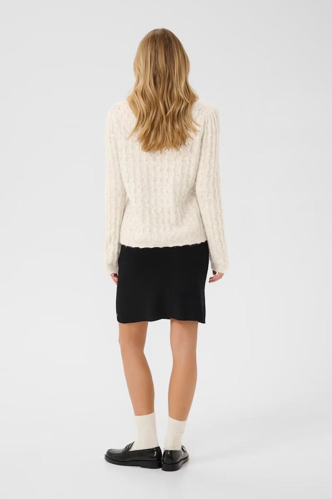 Femme vue de dos portant un chandail à maille torsadée beige avec une jupe noire et des chaussettes blanches.