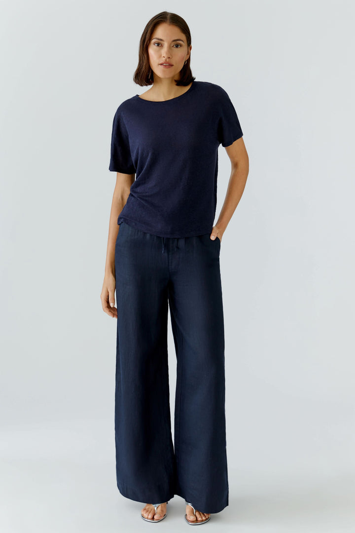 Femme portant un chandail à manches courtes fluide avec pantalon ample, style moderne et confortable, fond blanc.