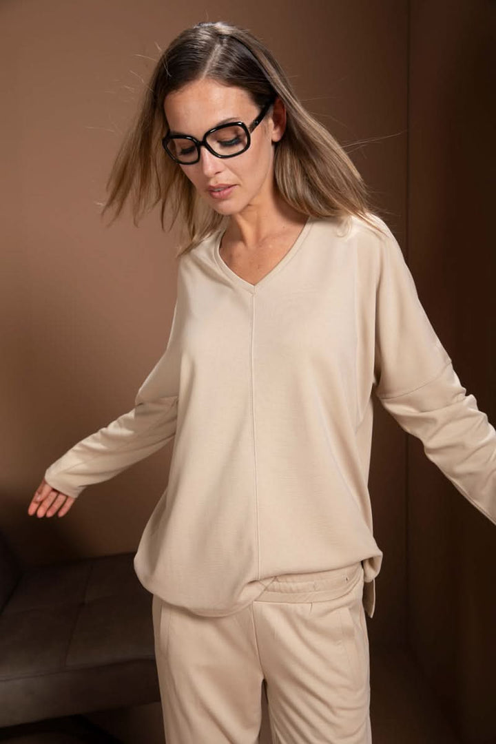 Femme portant un chandail minimaliste beige avec encolure en V, manches longues et épaules tombantes, look moderne et décontracté.