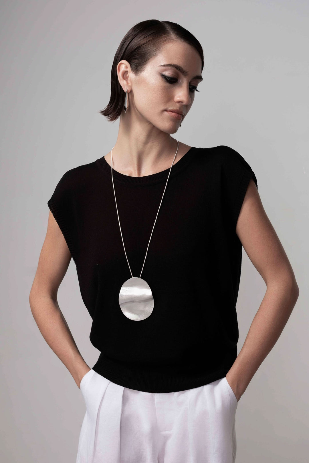 Femme portant un chandail ouverture au dos, sans manches, maille fine, avec un pendentif argenté, tenue élégante et moderne.