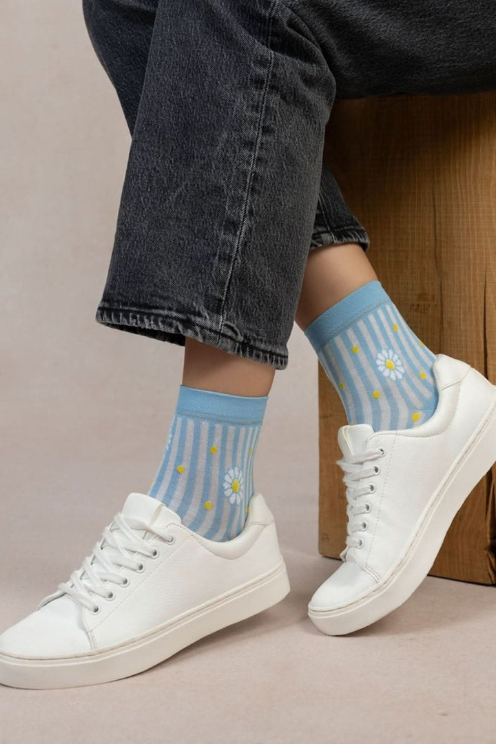 Chaussettes courtes à rayures bleues et marguerites, portées avec des chaussures blanches sur un fond neutre.