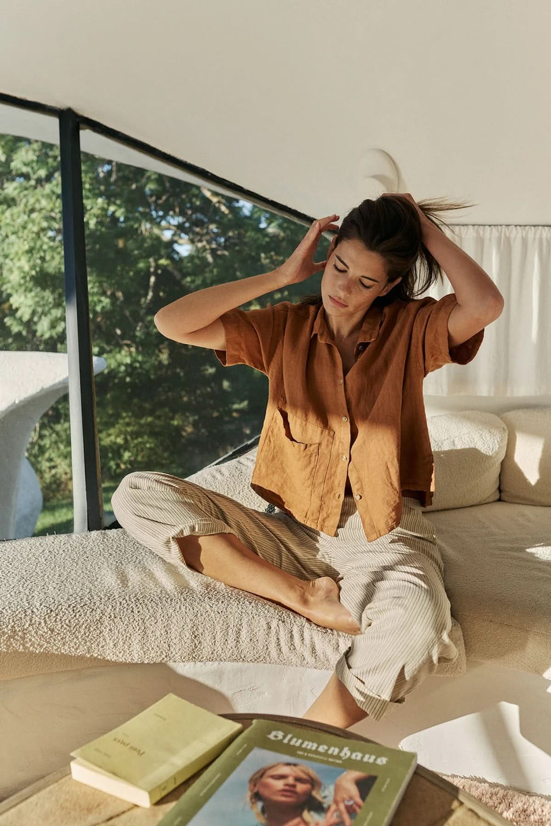 Femme portant une chemise en lin à coupe décontractée assise confortablement sur un lit, lumière naturelle environnante.
