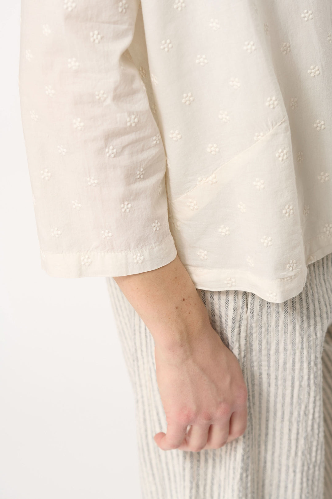 Détail d'une chemise sans col en coton léger brodé, portée avec un pantalon rayé.