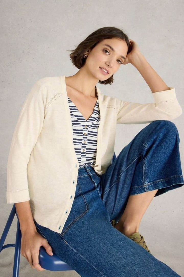 Femme portant un gilet Col V Naria blanc cassé, assise sur une chaise, avec un jean décontracté et un top rayé.