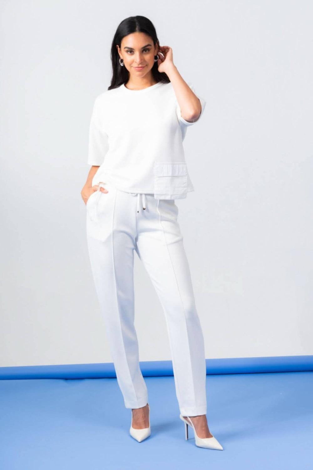 Haut à manches courtes blanc avec poche, porté avec un pantalon blanc et des chaussures à talons, sur fond bleu.