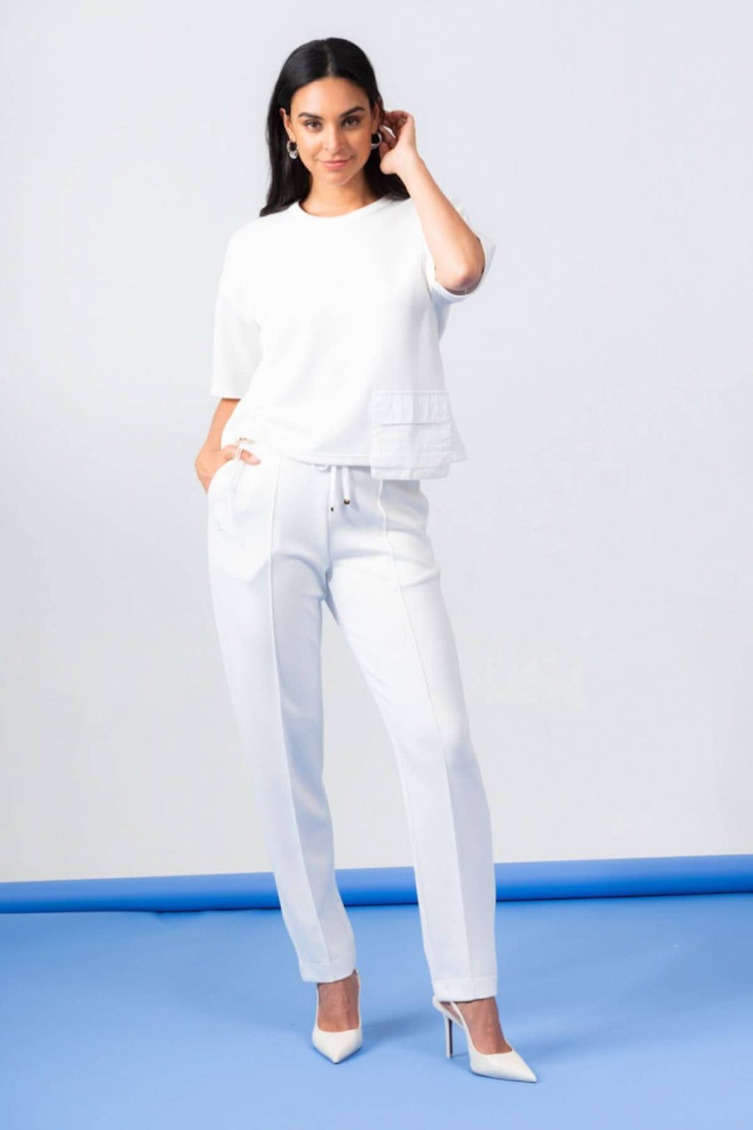 Haut à manches courtes blanc avec poche, porté avec un pantalon blanc et des chaussures à talons, sur fond bleu.