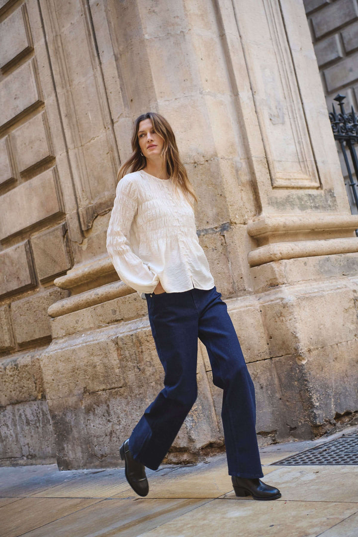 Femme portant un Jean Simona à jambe droite avec une blouse blanche, marchant dans un décor urbain élégant.