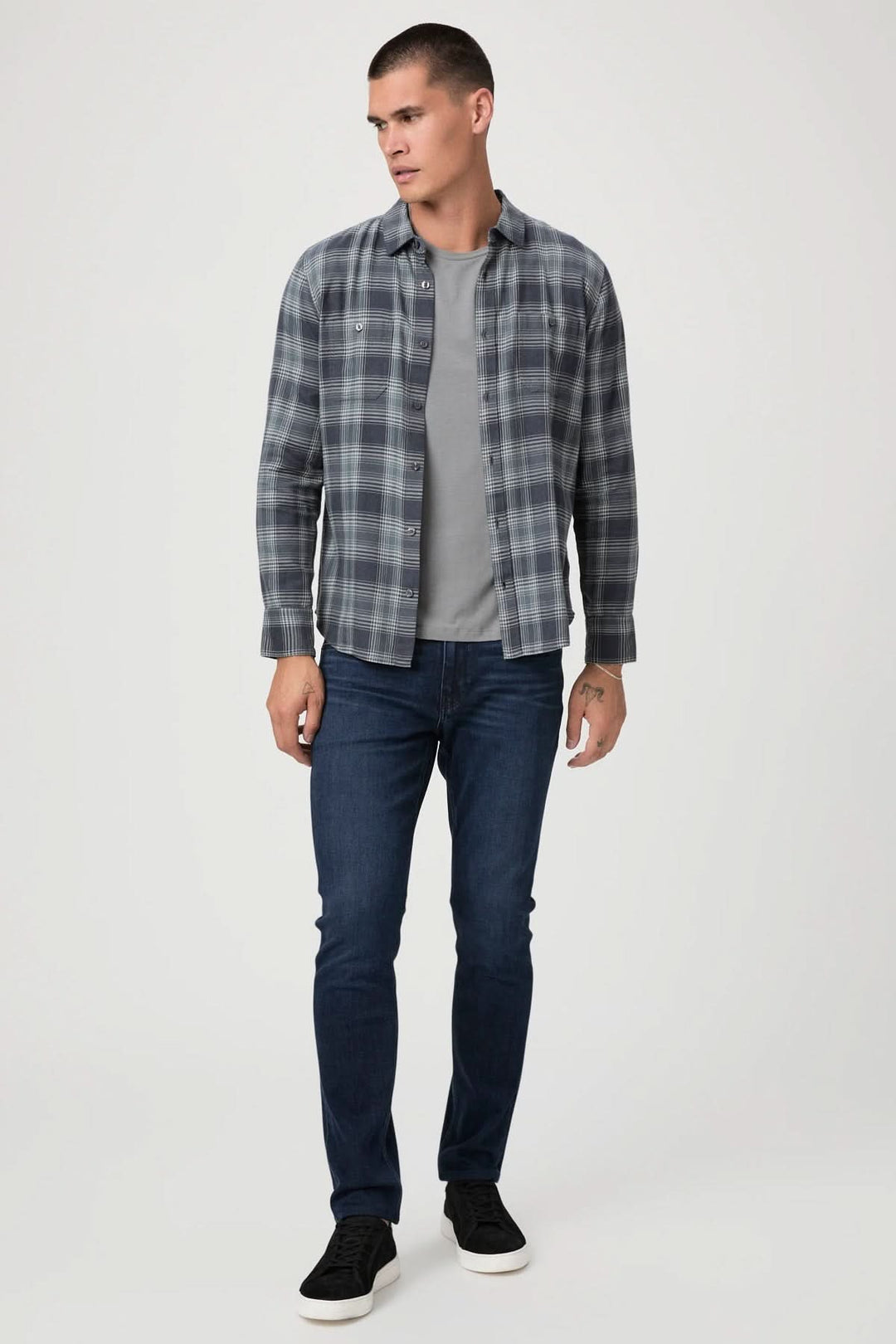 Homme portant une chemise à carreaux grise et un t-shirt, associé à un jean ajusté bleu et des baskets noires.