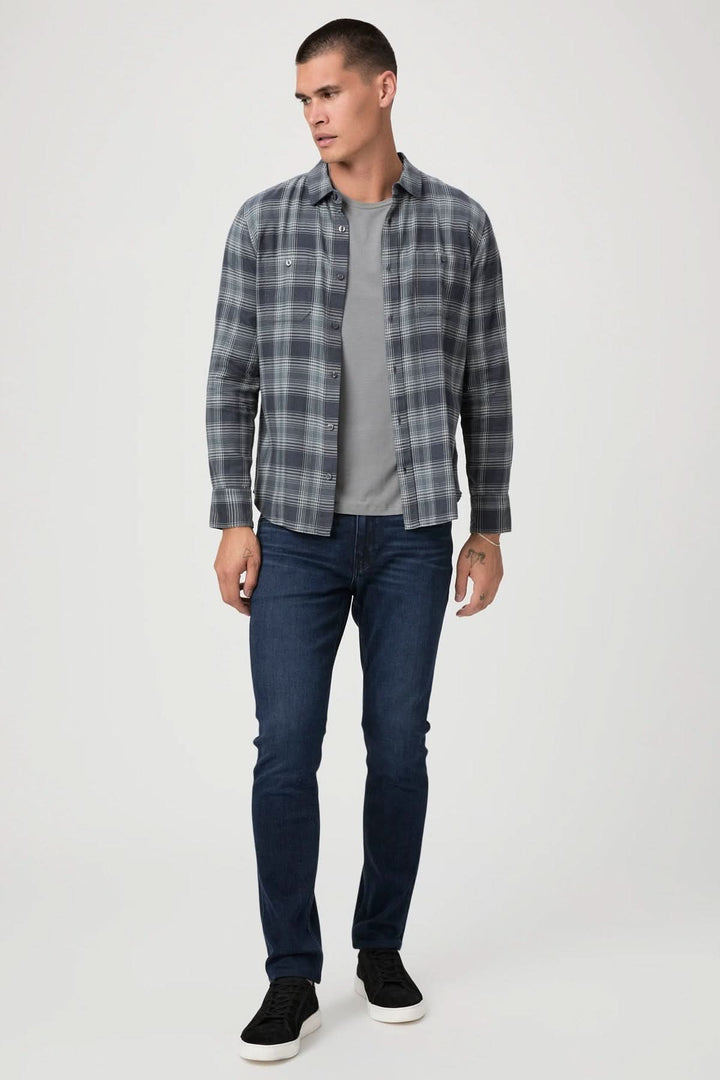 Homme portant une chemise à carreaux grise et un t-shirt, associé à un jean ajusté bleu et des baskets noires.