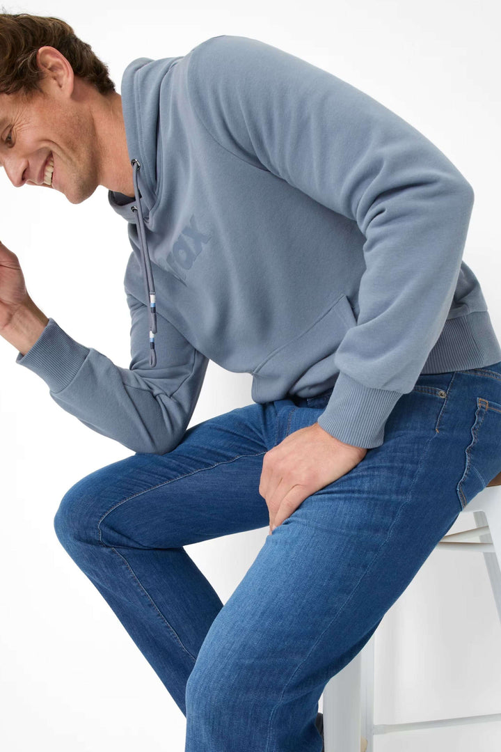 Homme souriant portant un sweat à capuche gris et un jean bleu élégant, assis sur une chaise.