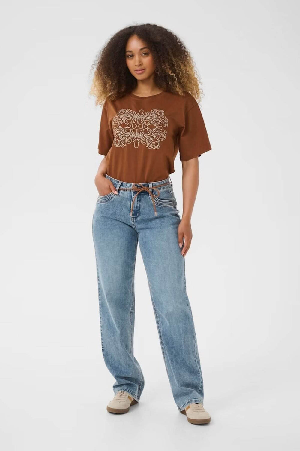 Femme portant un jean à coupe droite légèrement délavé, associé à un t-shirt graphique et des sneakers beige.