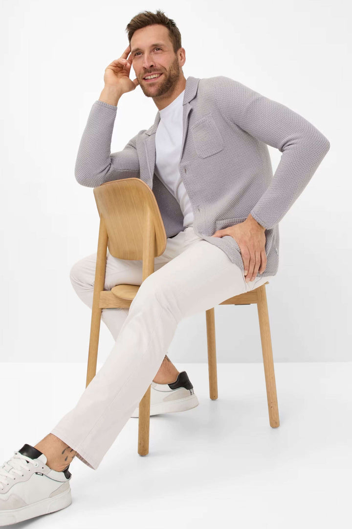 Homme assis sur une chaise, portant un cardigan gris clair, T-shirt blanc et pantalon beige, avec des baskets blanches.